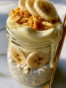 A handheld smartphone photo of a clear glass mason jar filled with high-protein banana pudding overnight oats and a gold spoon.