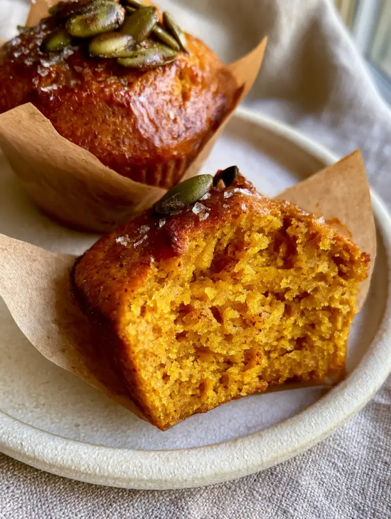 Close-up of golden-brown paleo pumpkin protein muffins topped with sea salt and pepitas on a stoneware plate.