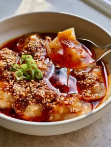 Handheld photo of a silver spoon lifting a chewy potato dumpling from a bowl of spicy potato dumplings with trailing red oil.